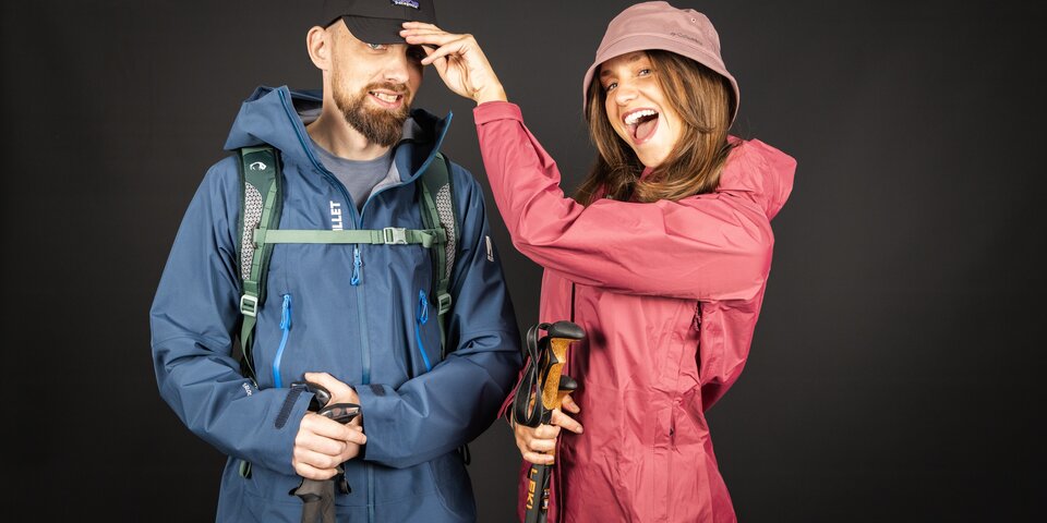 A happy couple dressed in hiking gear playfully poses against a dark background. © Sport Kessler GmbH