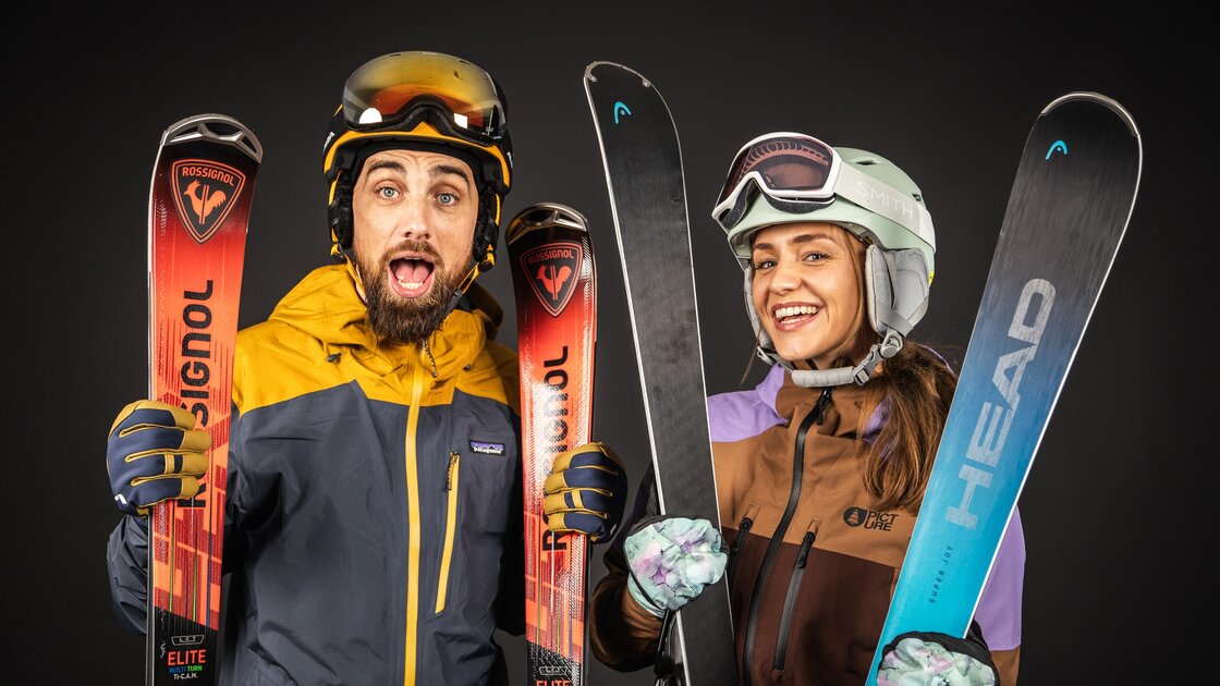 A man and a woman, both wearing ski gear, stand side-by-side, holding their skis. They appear happy and excited. The man has a beard and is wearing a yellow and dark blue jacket, while the woman has long brown hair and a brown and purple jacket. Their skis are different brands: the man's are Rossignol, and the woman's are Head. The overall impression is one of winter sports enthusiasm and readiness for the slopes. © Sport Kessler GmbH